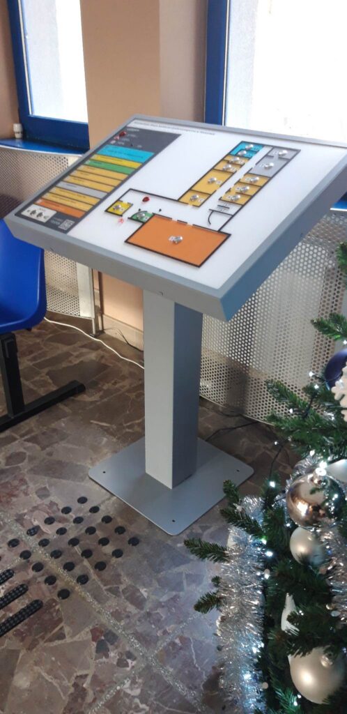 Interaktywna, dotykowa mapa tyflograficzna budynku z opisami w brajlu, oznaczeniami pomieszczeń i ścieżkami prowadzącymi.