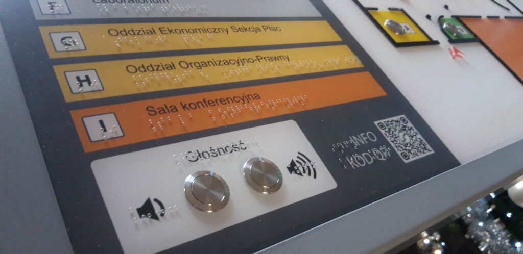 Tablica informacyjna z oznaczeniami pomieszczeń, pismem Braille’a, przyciskami dostępności i kodem QR.
