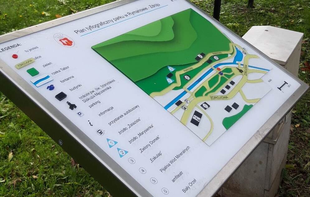 Tyflograficzny plan parku w Rymanowie-Zdroju z wypukłą mapą terenu, legendą, oznaczeniami dróg, rzeki, budynków, parkingu i punktów orientacyjnych.