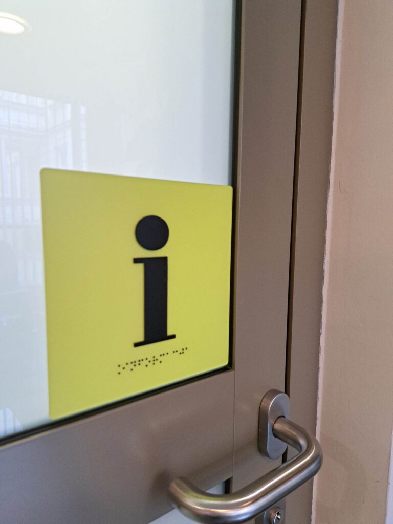 Tabliczka informacyjna z symbolem informacji „i” i napisem w alfabecie Braille’a.