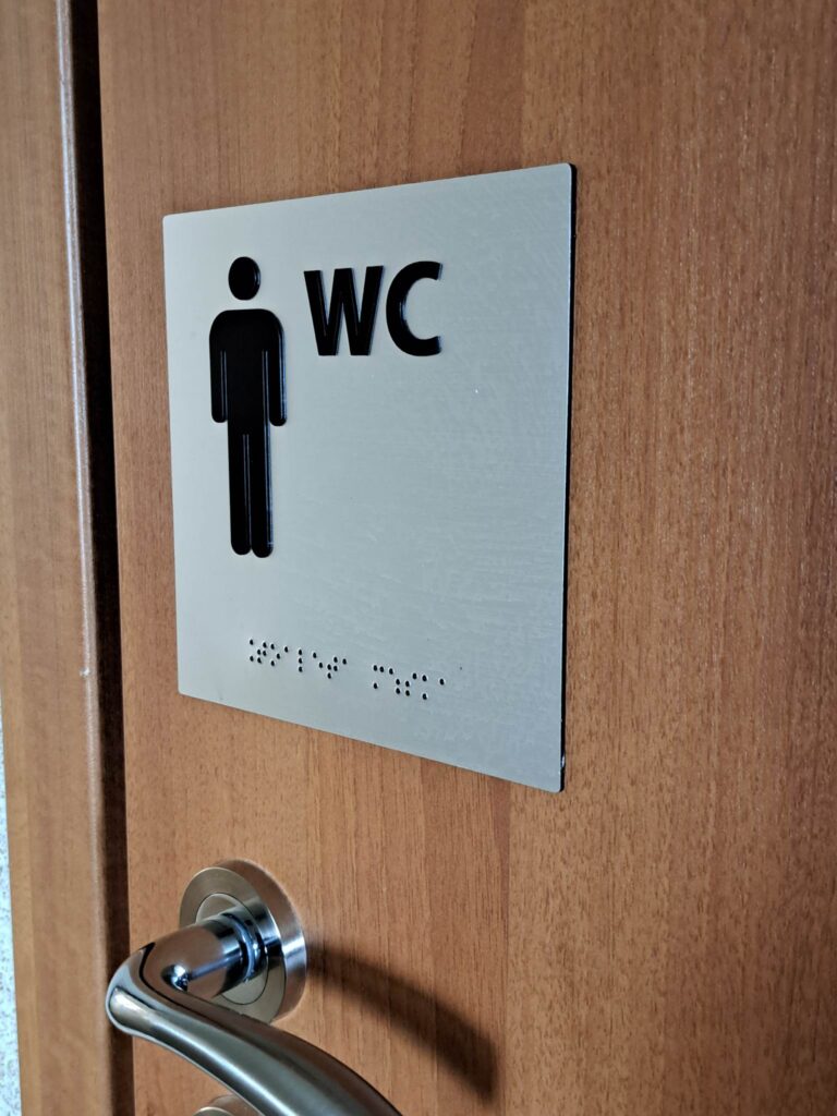 Tabliczka toalety z symbolem osoby, napisem „WC” i oznaczeniem w brajlu.