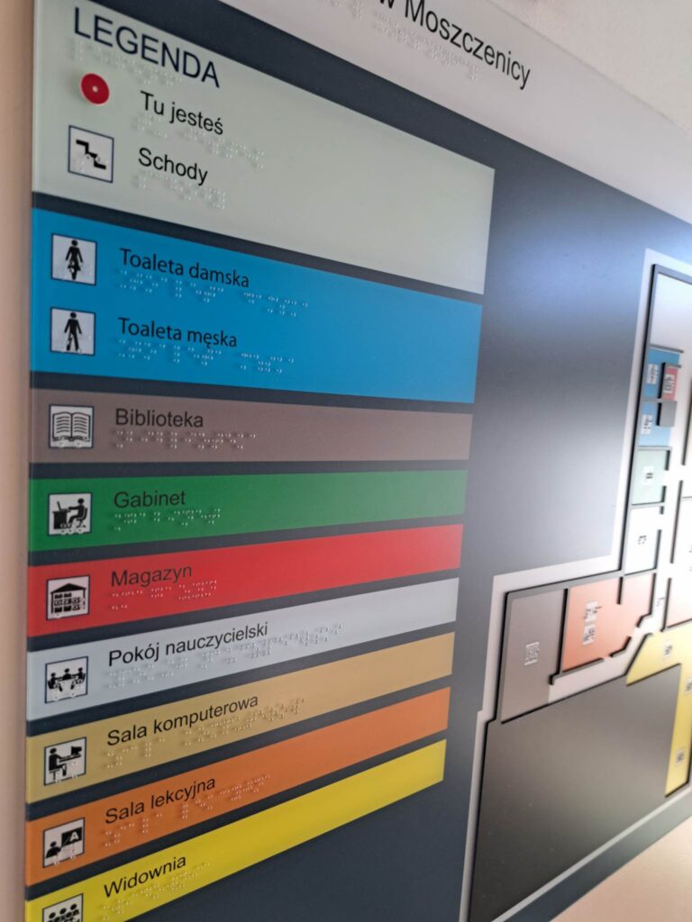 Tablica informacyjna z legendą oznaczeń, piktogramami i opisami w brajlu, przedstawiająca m.in. toalety, bibliotekę, gabinet, magazyn, pokój nauczycielski, sale oraz widownię.