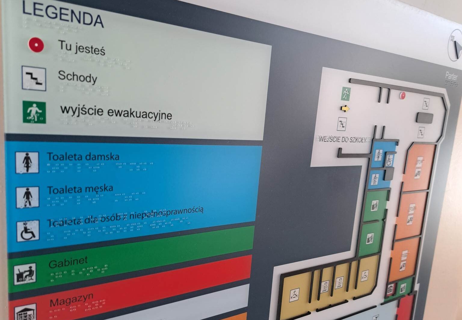 Tablica informacyjna z legendą oznaczeń dostępności i planem parteru budynku, z opisami toalet, gabinetu, magazynu, wyjść oraz schodów, uzupełniona o napisy Braille’a.
