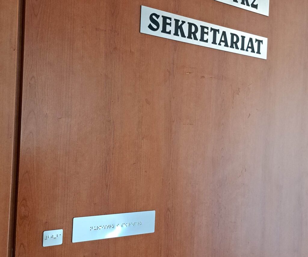 Tabliczka z napisem „SEKRETARIAT” oraz oznaczeniem brajlowskim na drzwiach.