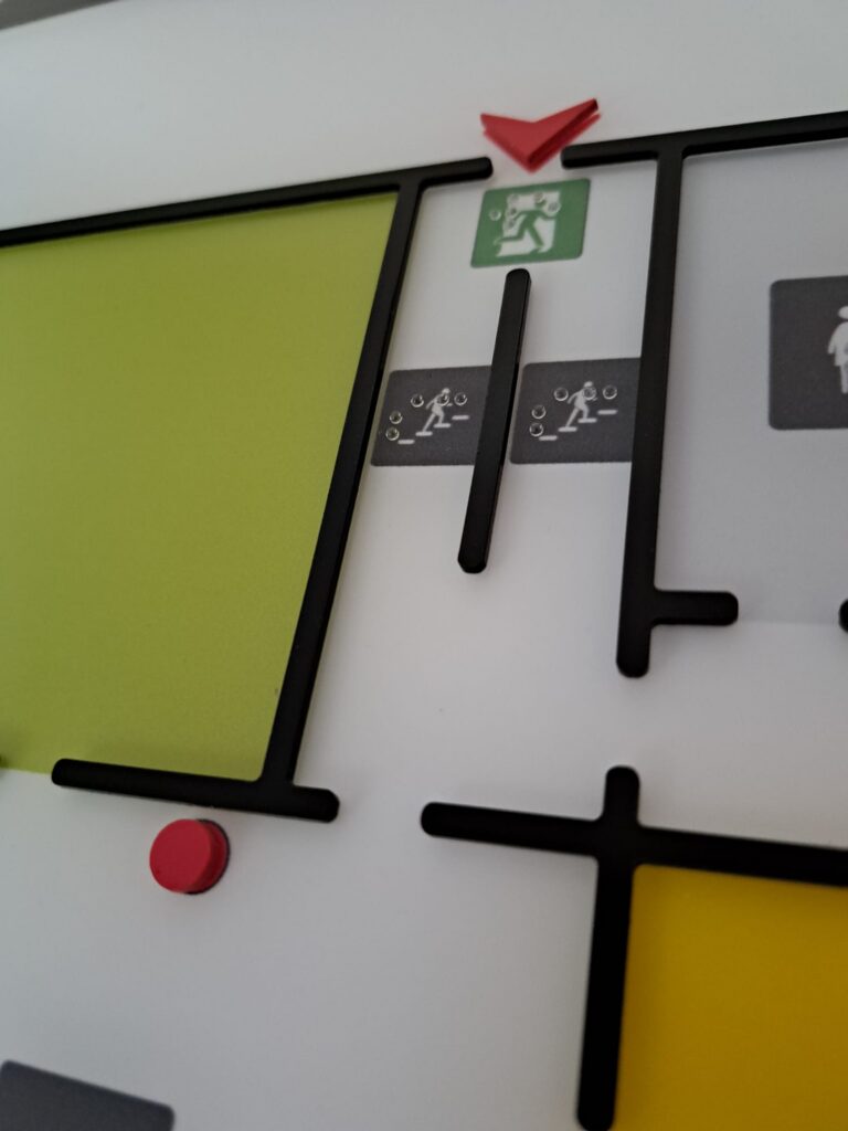 Fragment tyfloplanu z oznaczeniem wyjścia ewakuacyjnego, schodów i punktu „Tu jesteś”, z opisami w alfabecie Braille’a.