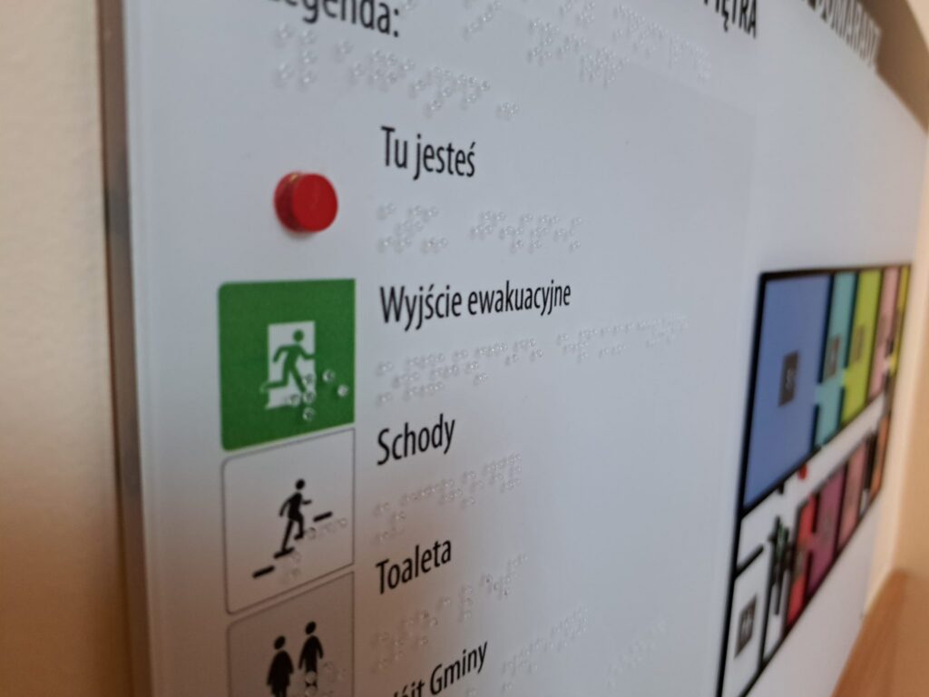 Zbliżenie legendy tyfloplanu z oznaczeniami „Tu jesteś”, wyjścia ewakuacyjnego, schodów i toalety, uzupełnionymi o opisy w alfabecie Braille’a.