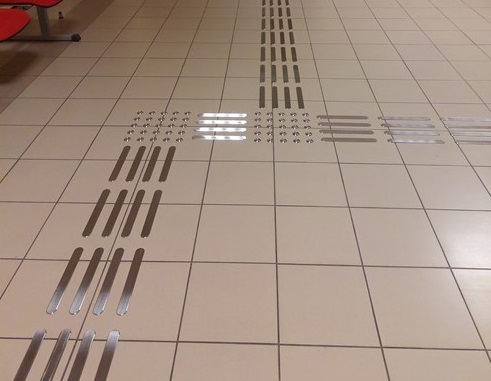 Ścieżka dotykowa z metalowych elementów na posadzce, prowadząca przez korytarz i wyposażona w oznaczenia kierunku oraz pola uwagi.