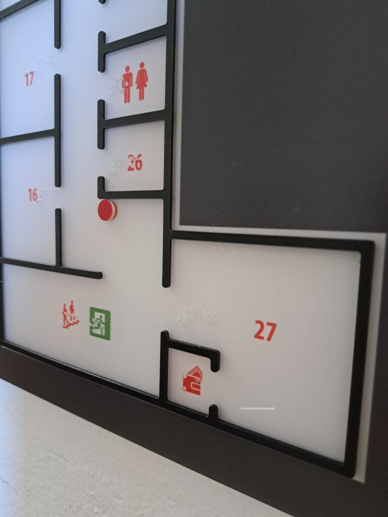 Zbliżenie na fragment tyflograficznego planu budynku z numerami pomieszczeń, symbolami toalet i wyjścia ewakuacyjnego oraz oznaczeniami w alfabecie Braille’a.