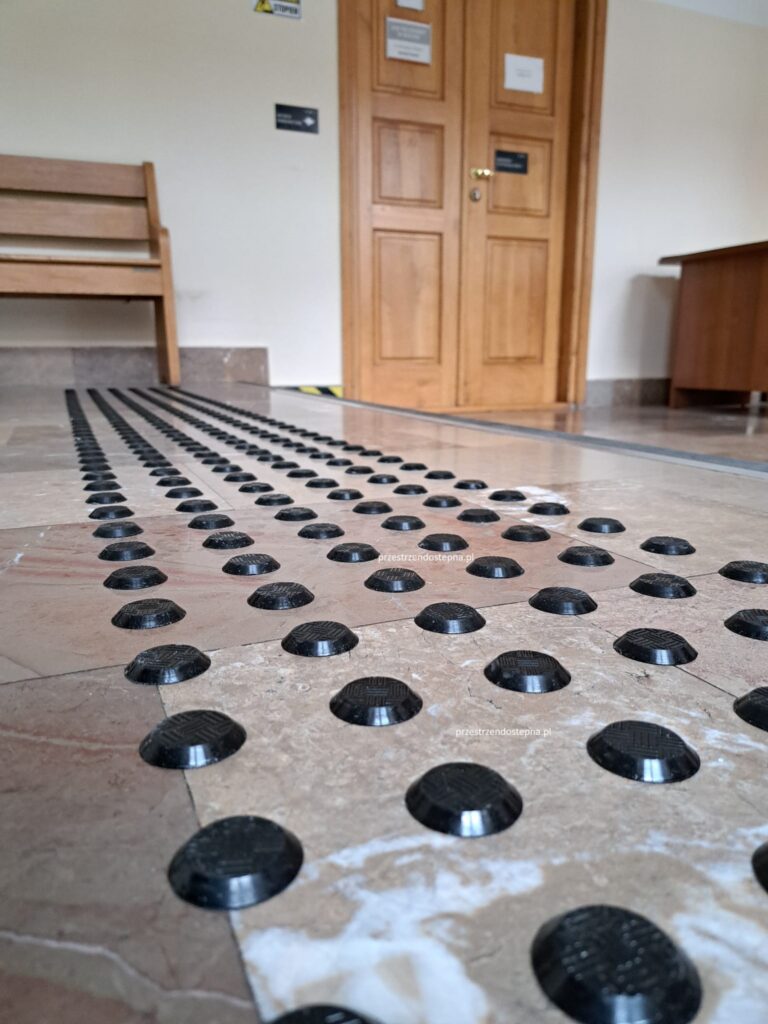 Ścieżka dotykowa z czarnymi wypukłymi punktami prowadząca wzdłuż korytarza.
