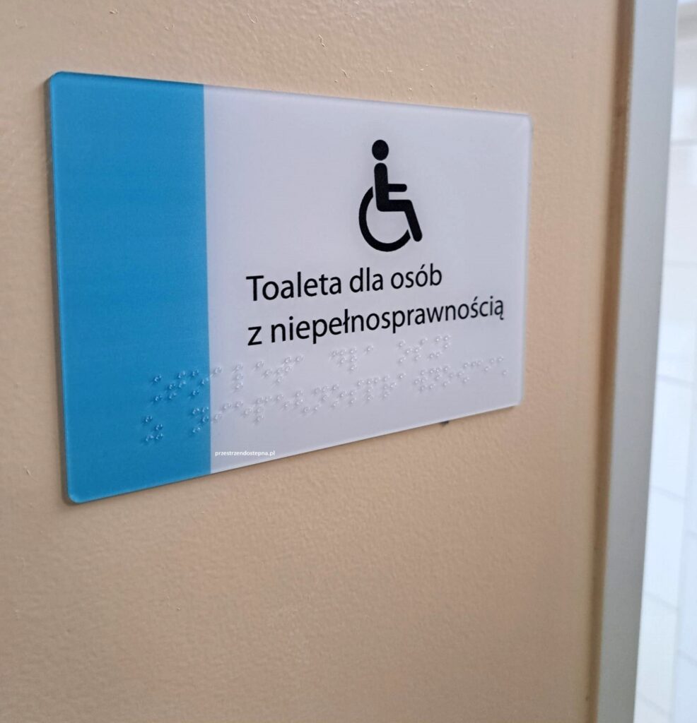 Tabliczka toalety dla osób z niepełnosprawnością z symbolem osoby na wózku i oznaczeniem w alfabecie Braille’a.