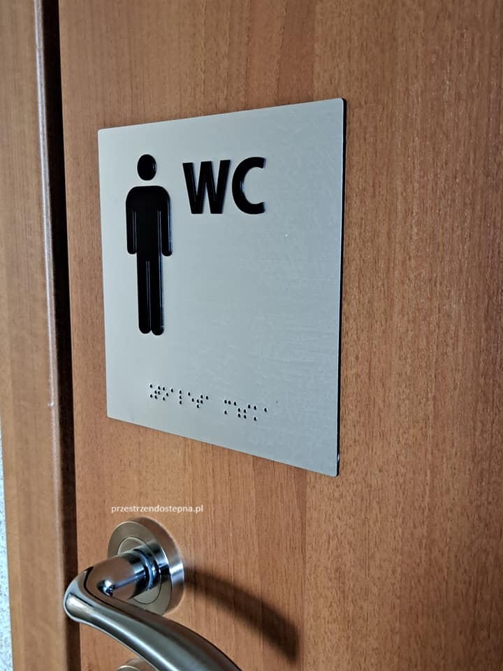 Tabliczka na drzwiach toalety z symbolem mężczyzny, oznaczeniem WC i napisem w alfabecie Braille’a.