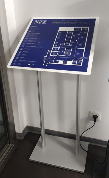 Stojakowa tablica informacyjna NFZ z planem pomieszczeń i oznaczeniami dotykowymi.