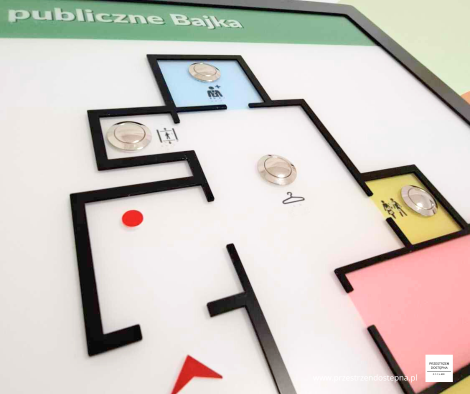 Dotykowa mapa przedszkola z wypukłym planem, oznaczeniami pomieszczeń i czerwonym punktem lokalizacji.