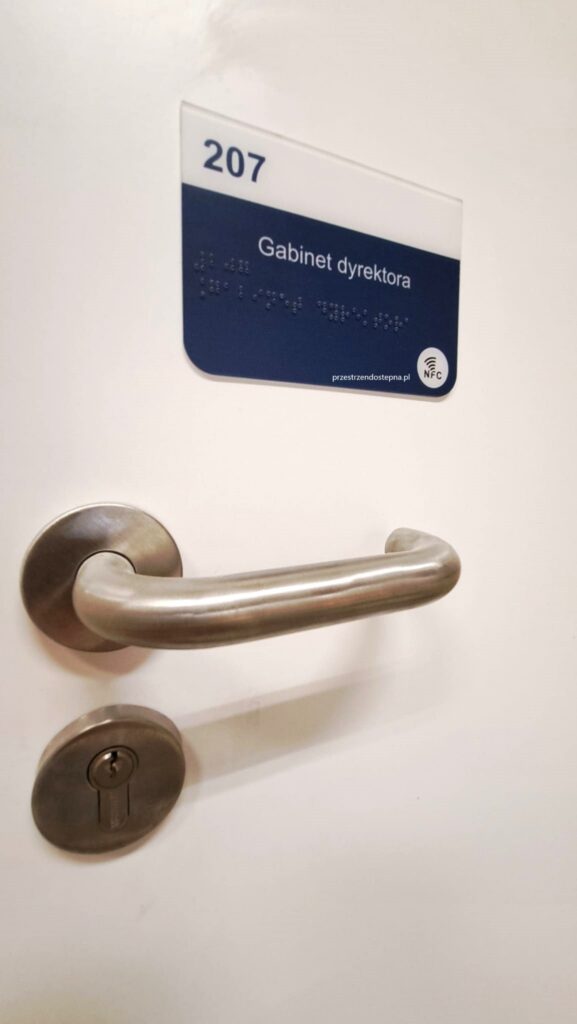Tabliczka drzwiowa gabinetu dyrektora, numer 207, z oznaczeniem w alfabecie Braille’a i symbolem NFC; poniżej widoczna klamka drzwi.